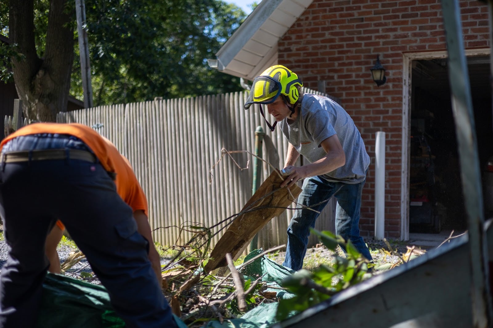 Two men in a yard, one wearing an orange shirt and the other a yellow hard hat, cleaning up yard waste.