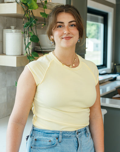 A young woman smiling in a kitchen with a plant. She is wearing a yellow top and jeans.
