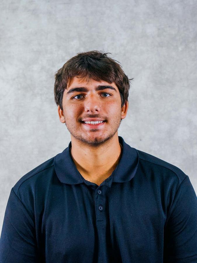 A smiling man with light facial hair wearing a dark blue polo shirt against a gray backdrop. The portrait is professionally lit for a clean and approachable look.
